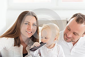 Parents sitting on the sofa. Their beautiful baby girl playing with cell phone.