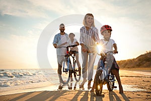 Parents teaching children to ride bicycles on sandy beach near sea at sunset