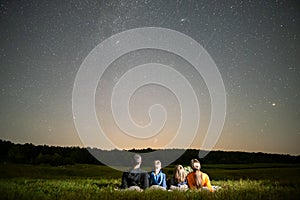 Parents and their kids resting in night field observing dark sky with many bright stars