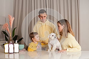 Parents in yellow sweaters, mother and father with child son and white dog, three abstract plastic bottles on the table