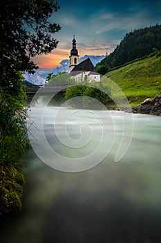 The parish church of St. Sebastian in Ramsau near Berchtesgaden as a long exposure with the river and a bridge at sunset