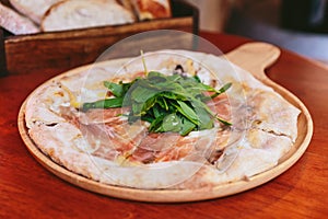 Parma Ham Pizza topping with rocket on rounded wooden plate with sliced bread in wooden box in the background