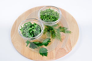 Parsley and dill leaves in two glass bowls and lone twigs on round wooden cutting board
