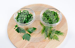 Parsley and dill leaves in two glass bowls and lone twigs on round wooden cutting board view from above