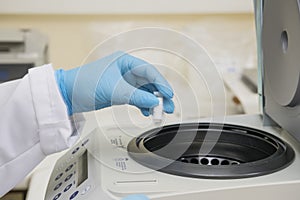 A part of an arm with labcoat and with blue nitril gloves putting a small plastic test tube in a micro centrifuge.