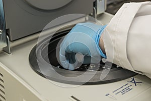 A part of an arm with labcoat and with blue nitril gloves putting a small plastic test tube in a micro centrifuge.