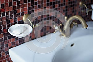 Part of bath room - wash basin with bronze crane, white rack for soap, brown mosaic tile background. Blurred picture.