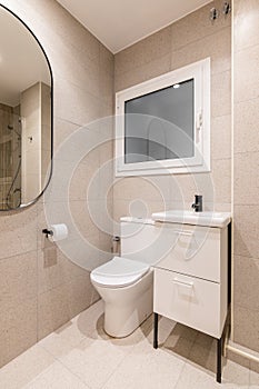 Part of bathroom with toilet bowl, washbasin on table, beautiful oval mirror on wall lined with beige marble tiles, in
