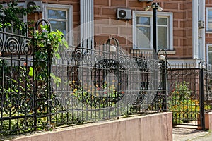 Part of a black fence made of iron bars on a concrete foundation and an open gate at the brown wall