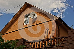 Part of the brown house behind the fence with a window in the attic and a satellite dish on the wall