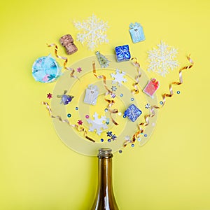 Part of champagne party bottle with confetti of gifts, sequins, ribbons, snowflakes on bright yellow background. Flat lay. Celebra