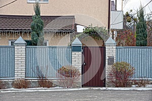 Part of a fence of gray metal and brick with a closed brown door on a winter street