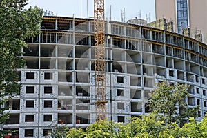 Part of a large unfinished gray building with a tower crane in the street with green tree branches