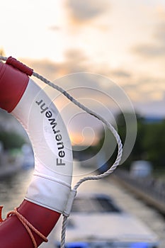 Part of a lifebuoy with rope. In the blurry background you can see a river on which a boat is driving, and the sky lit by the even