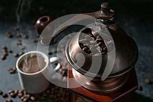 Part of a manual vintage coffee grinder, cup with the hot drink and roasted beans on a dark rustic background, selected focus