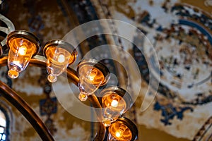 Part of a modern electric chandelier with warm light in front of a blurred painted ceiling in a historic building.