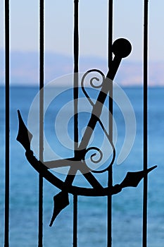 Part of wrought iron fence with silhouette of old antique vintage handmade anchor with decorations and light blue sea in back