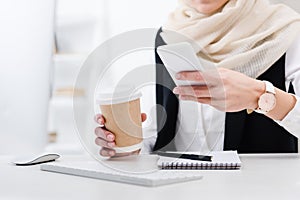 partial view of businesswoman with coffee to go using smartphone