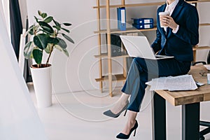 partial view of businesswoman with laptop and coffee to go sitting on table