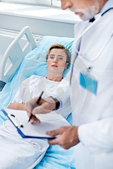 partial view of male doctor with stethoscope over neck writing in clipboard near adult female patient in hospital