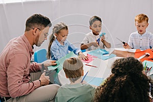 partial view of male teacher and multiracial preschoolers cutting colorful papers with scissors at table