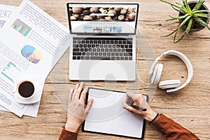 partial view of man working at laptop with depositphotos website, headphones, textbook, pen, infographics, coffee cup and potted