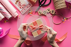 Partial view of woman decorating gift box with ribbon near valentines handiwork supplies on pink background