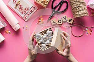 Partial view of woman holding gift box with silver hearts near valentines handiwork supplies on pink background