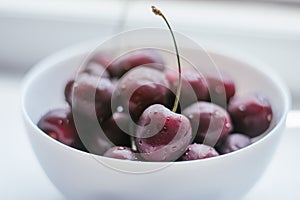 Partially blurred white bowl with ripe red sweet cherries, on white surface