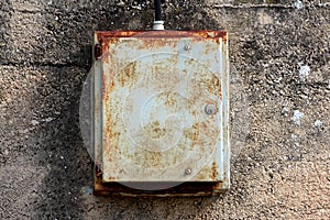 Partially rusted dilapidated locked metal electrical box mounted on cracked aged concrete wall connected with thick black