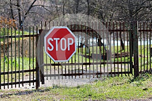 Partially rusted metal locked picket fence driveway doors with large stop road sign mounted in front surrounded with uncut grass