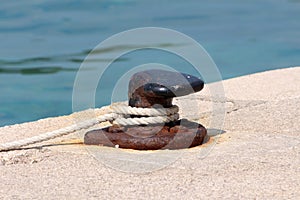Partially rusted strong old iron mooring bollard screwed on local stone pier with five rusted metal bolts with white rope tied