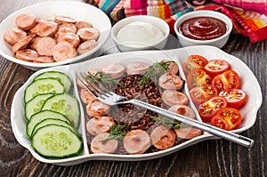 Partitioned dish with fried sausages, red rice, cucumbers, tomato, fork, ketchup and mayonnaise, napkin on table