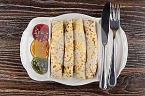 Partitioned dish with jams, pancake rolls, fork and knife on table. Top view