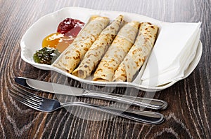 Partitioned dish with jams, pancake rolls, tissue, fork, knife on wooden table