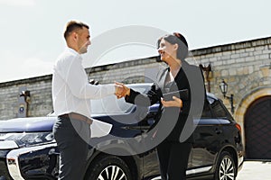 Partners, greeting. Young adult smiling man and woman in business dark suits standing near a car, shaking hands