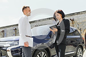 Partners, greeting. Young adult smiling man and woman in business dark suits standing near a car, shaking hands