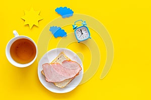 Parts of day. Morning. Time for breakfast. Tea, sandwich near alarm clock, sun and clouds cutout on yellow background