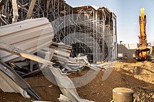 Parts of the ruins of a partially demolished high-bay warehouse with a demolition excavator in the background