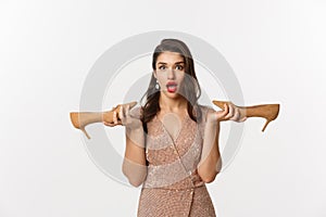 Party and celebration concept. Attractive woman in elegant dress holding pair of heels and looking surprised, dressing