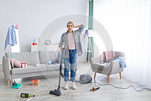 After party chaos. Dismayed millennial woman standing in her messy apartment with vacuum cleaner, holding head in terror