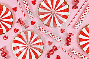 Party flat lay with red paper plates, heart ornaments, candy and drinking straws on pink  background