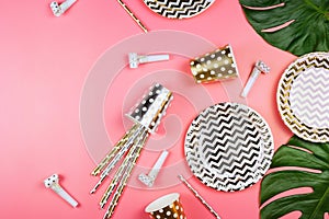 Party golden and silver paper glasses, dishes and straws, party horns and monstera leaves on a table.