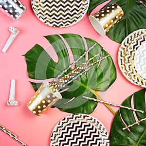 Party golden and silver paper glasses, dishes and straws, party horns and monstera leaves on a table.