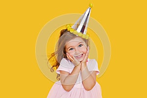 Party time. A joyful cute little child girl in a festive cap and elegant dress celebrates her birthday on a yellow