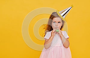 Party time. A joyful little girl in a festive cap and elegant dress celebrates her birthday. Blowing a whistle