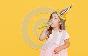 Party time. A joyful little girl in a festive cap and elegant dress celebrates her birthday. Blowing a whistle on a yellow