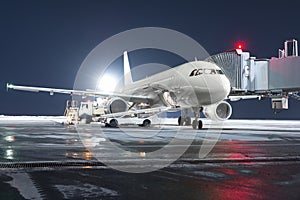 The passenger aircraft stands at the boarding bridge on night airport apron. The baggage compartment of the aircraft is open and
