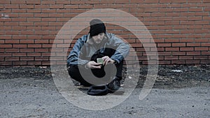 Passerby throws handout of 1 dollar money to homeless beggar. Refugee needs help. Tramp sits in dirty clothes, hat cap