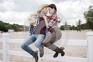 Passionate cowboy style couple kissing while sitting on fence at ranch